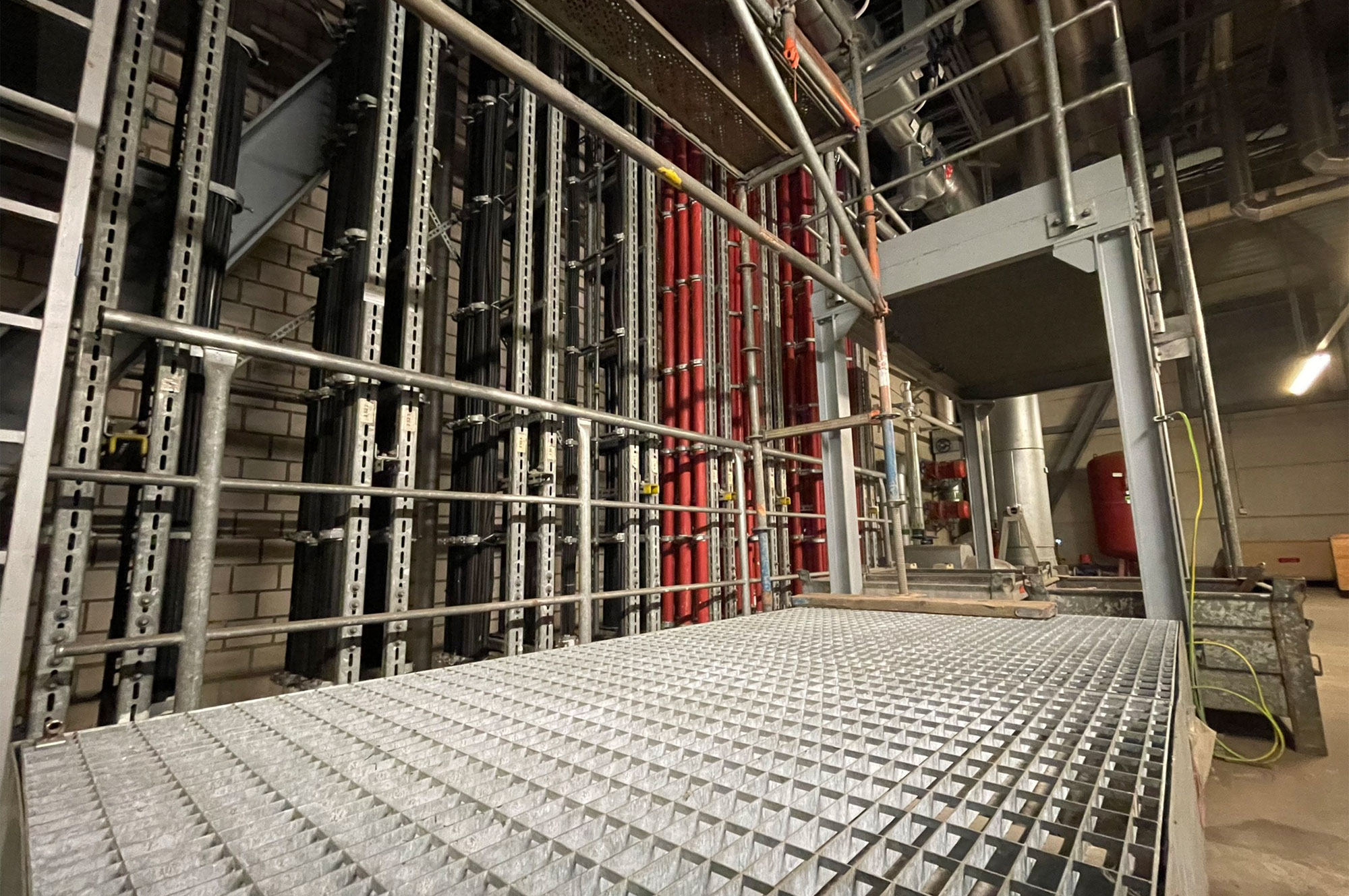 Metallgitterboden in einer industriellen Umgebung mit vertikalen Rohren in verschiedenen Farben im Hintergrund.