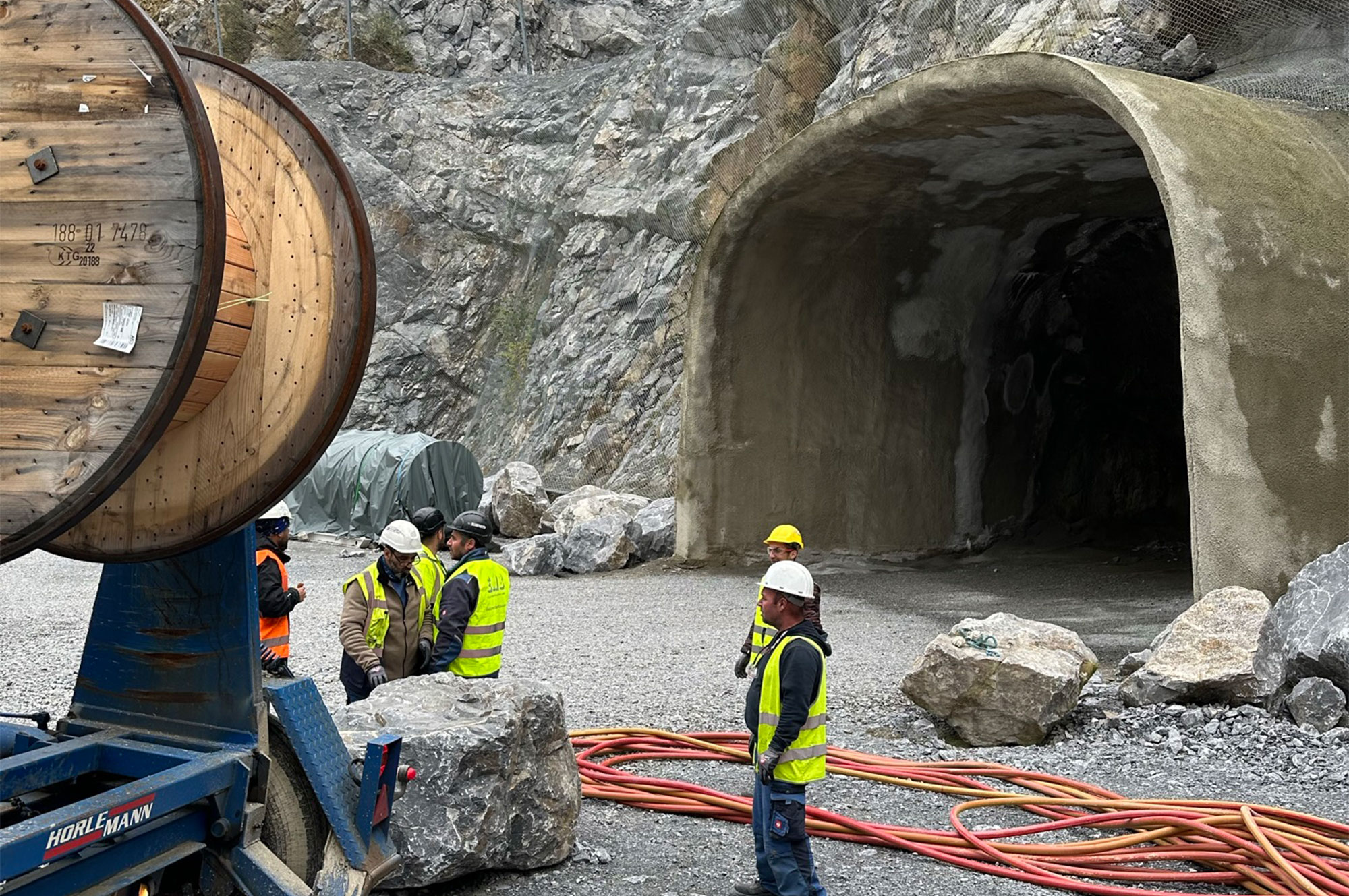Arbeiter in Sicherheitskleidung stehen auf einer Baustelle neben Kabeltrommeln und einem Tunnelportal in einem Felsbereich.