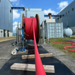 Rote Energiekabel auf einer großen Rolle, die auf einem Betonboden verlegt wird, mit industriellen Gebäuden im Hintergrund.