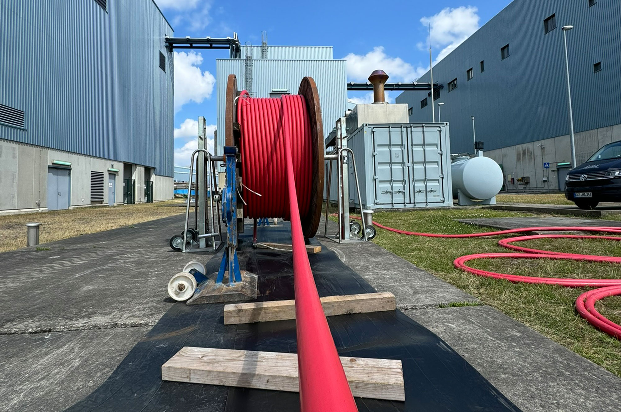 Rote Energiekabel auf einer großen Rolle, die auf einem Betonboden verlegt wird, mit industriellen Gebäuden im Hintergrund.