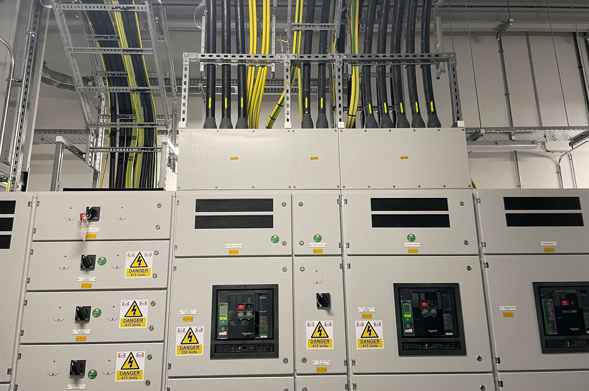Verteilerkästen mit Schaltern und Warnhinweisen in einem technischen Raum, Kabelsysteme verlaufen an der Decke.