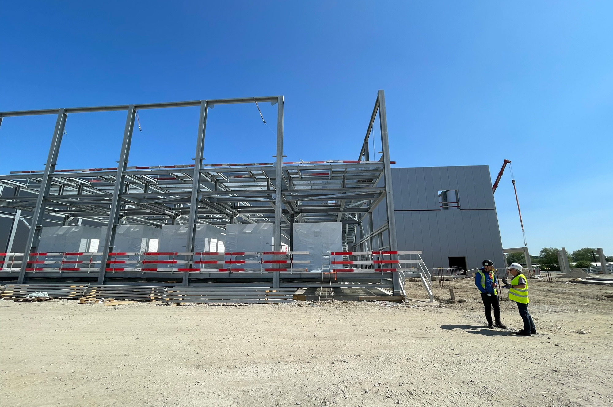 Bauarbeiten an einem modernen Gebäude mit Stahlrahmen und transparenten Wänden, zwei Personen in Sicherheitskleidung stehen davor.