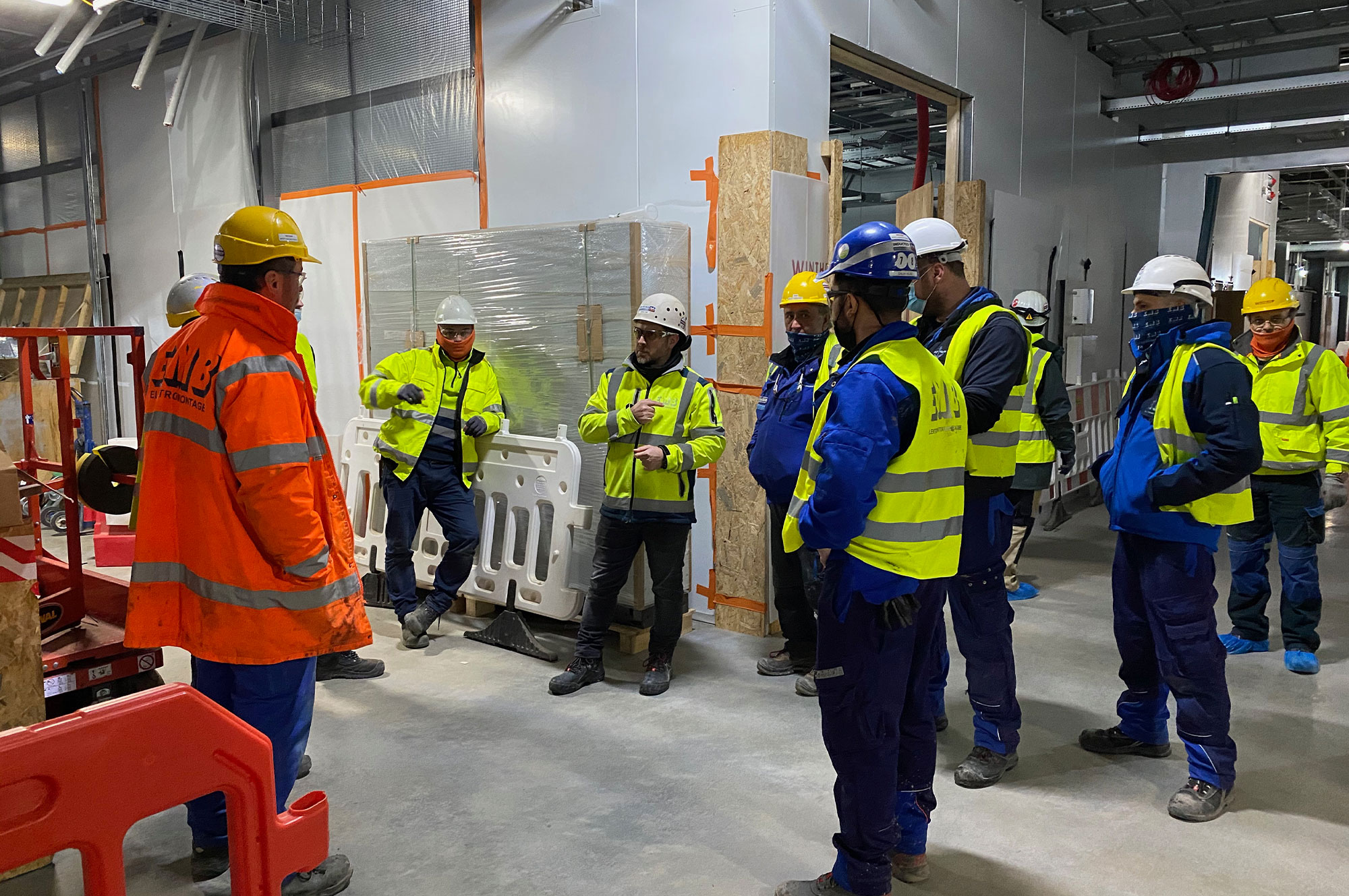 Gruppe von Bauarbeitern in Sicherheitskleidung und Helmen, die einem Vorgesetzten auf einer Baustelle zuhören.