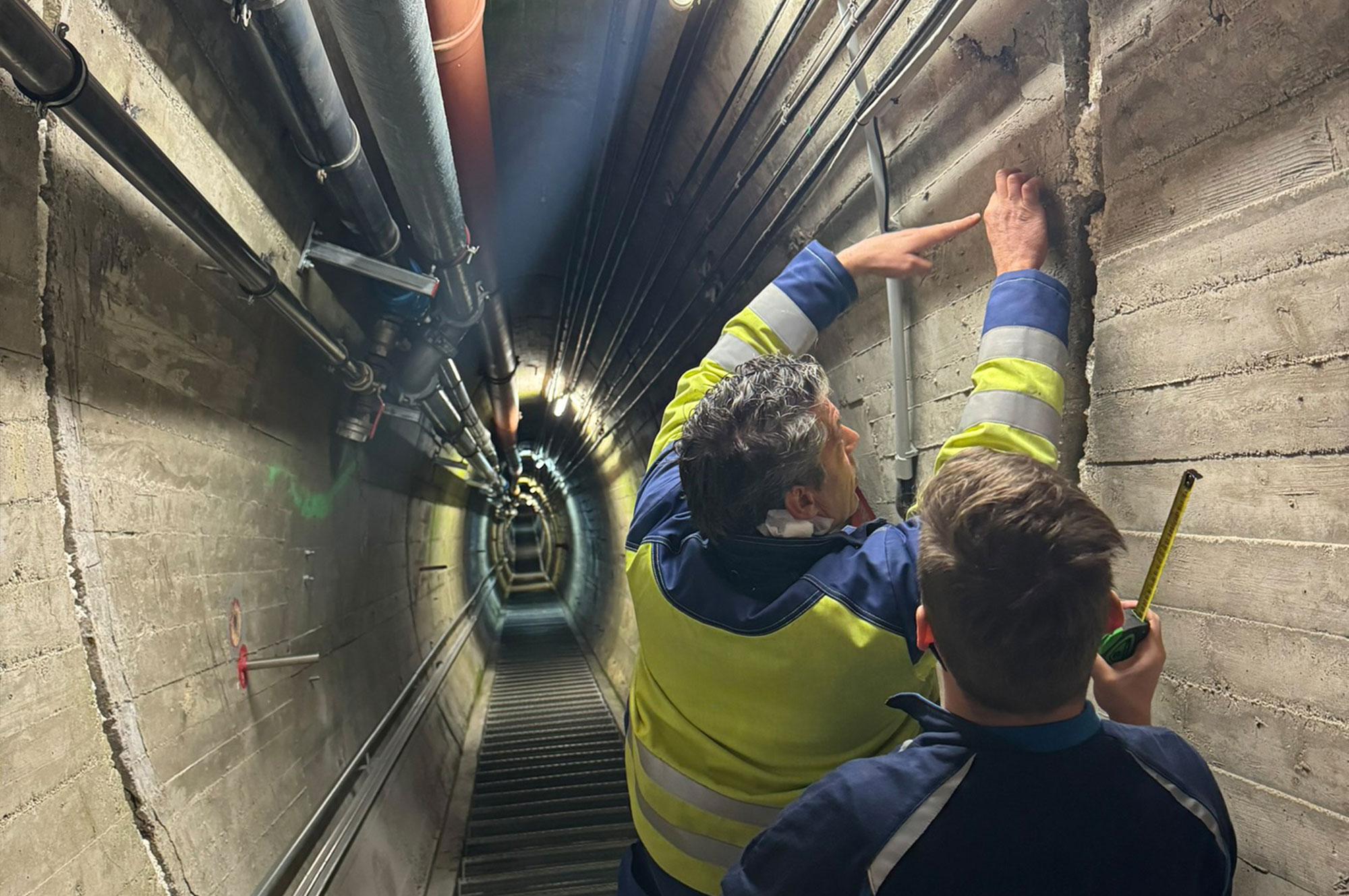 Zwei Personen in Arbeitskleidung untersuchen eine Wand in einem Tunnel, während sie Messwerkzeuge verwenden.