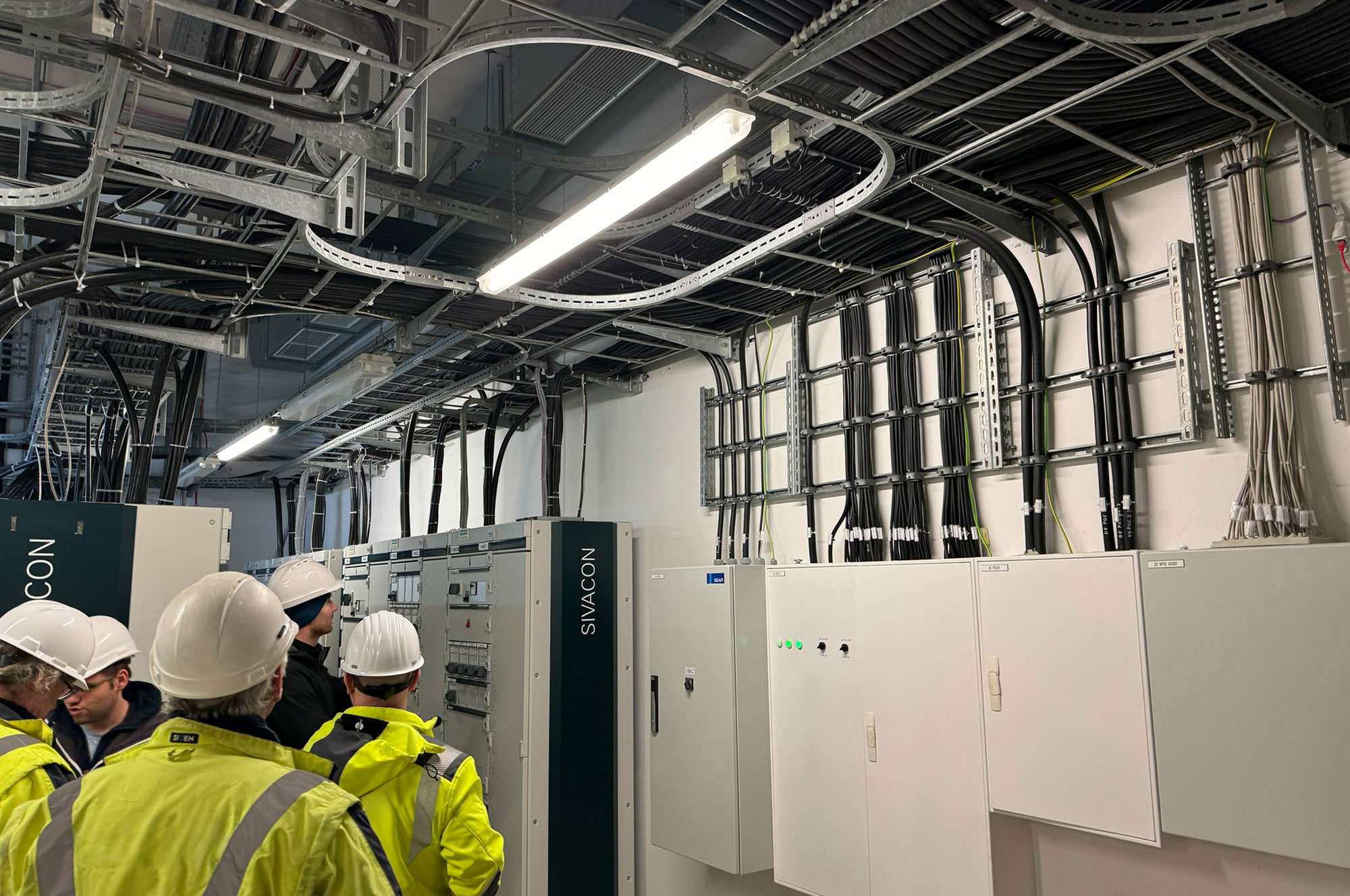 Techniker in Sicherheitshelmen und -jacken stehen vor elektrischen Schaltanlagen und Kabelinstallationen in einem technischen Raum.