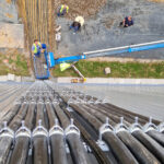Blick von oben auf eine Baustelle mit Arbeitern, die an Kabeln und einer Hebebühne arbeiten, die entlang einer vertikalen Struktur verlegt sind.