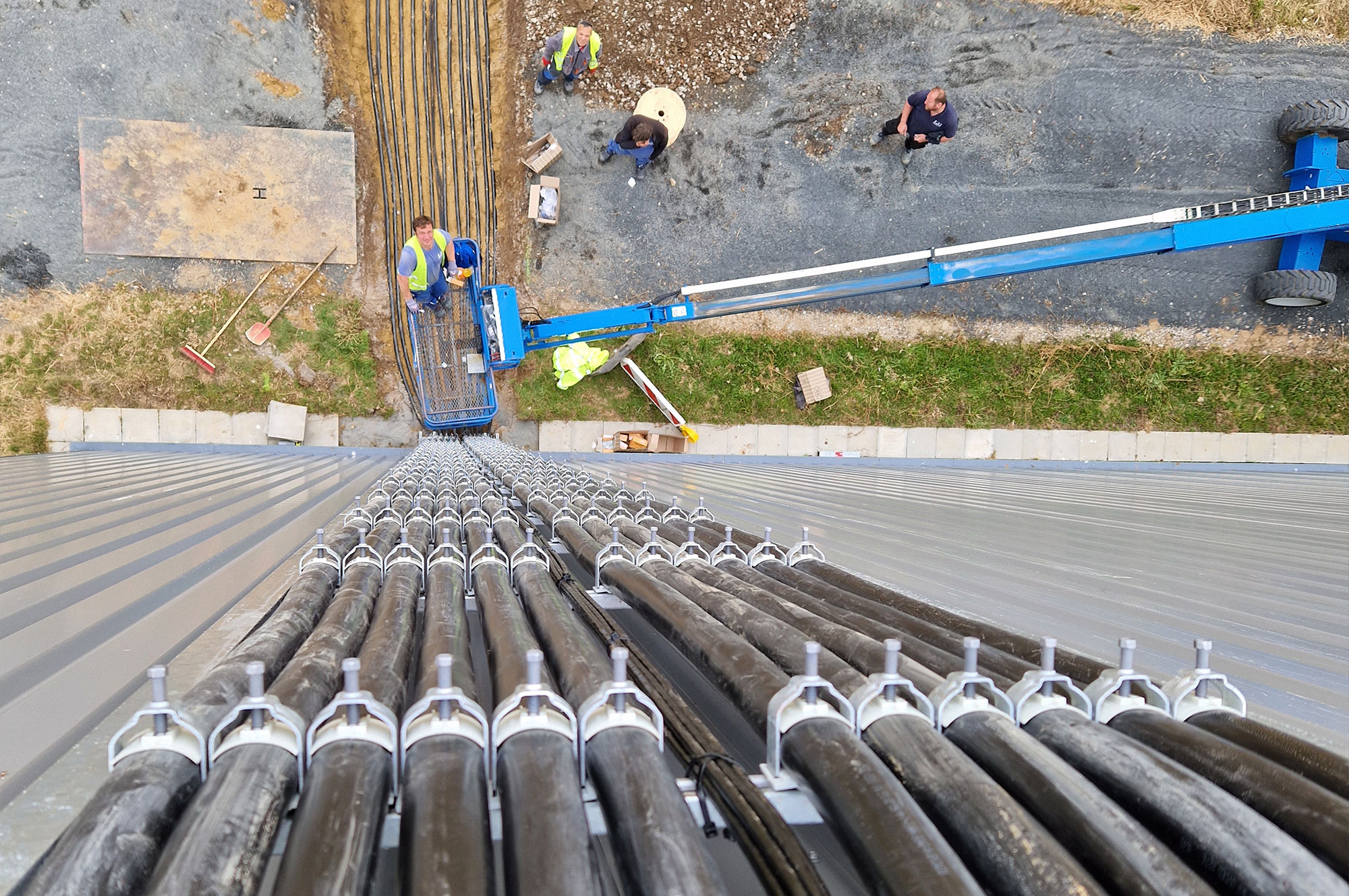 Blick von oben auf eine Baustelle mit Arbeitern, die an Kabeln und einer Hebebühne arbeiten, die entlang einer vertikalen Struktur verlegt sind.