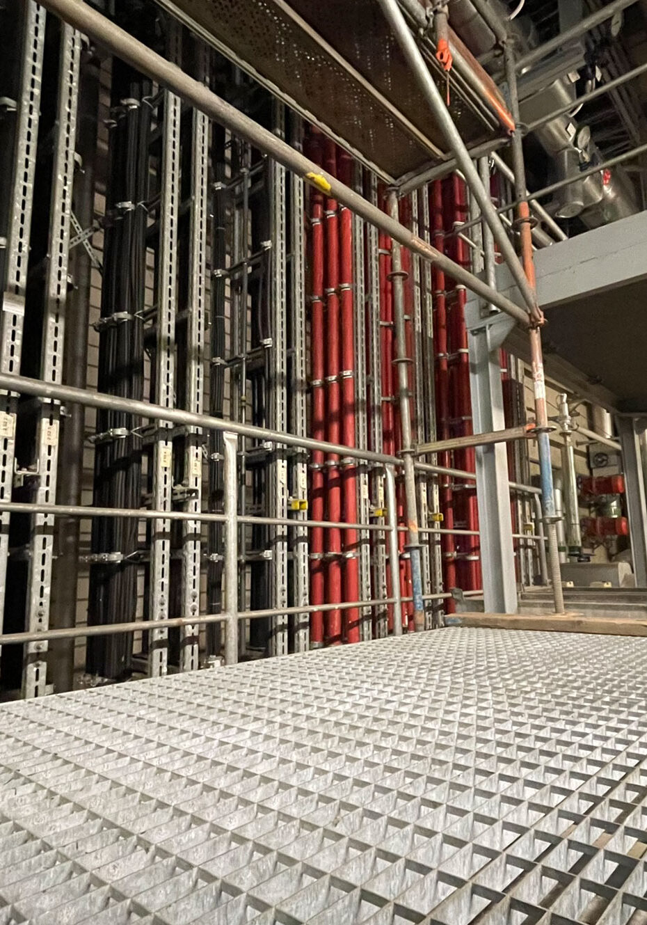 Metallgitterboden in einer industriellen Umgebung mit vertikalen Rohren in verschiedenen Farben im Hintergrund.