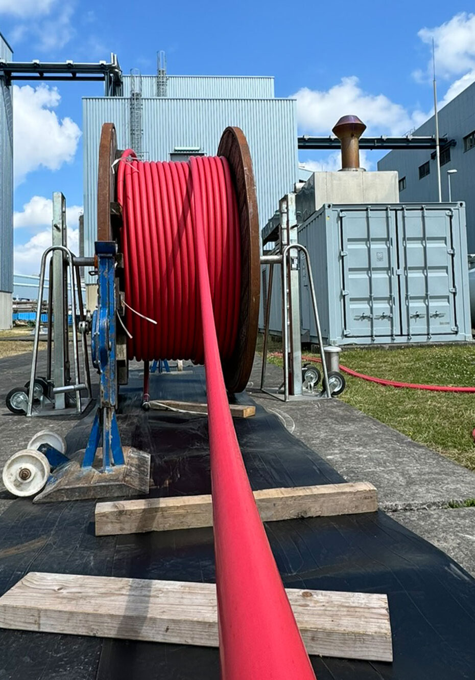 Rote Energiekabel auf einer großen Rolle, die auf einem Betonboden verlegt wird, mit industriellen Gebäuden im Hintergrund.