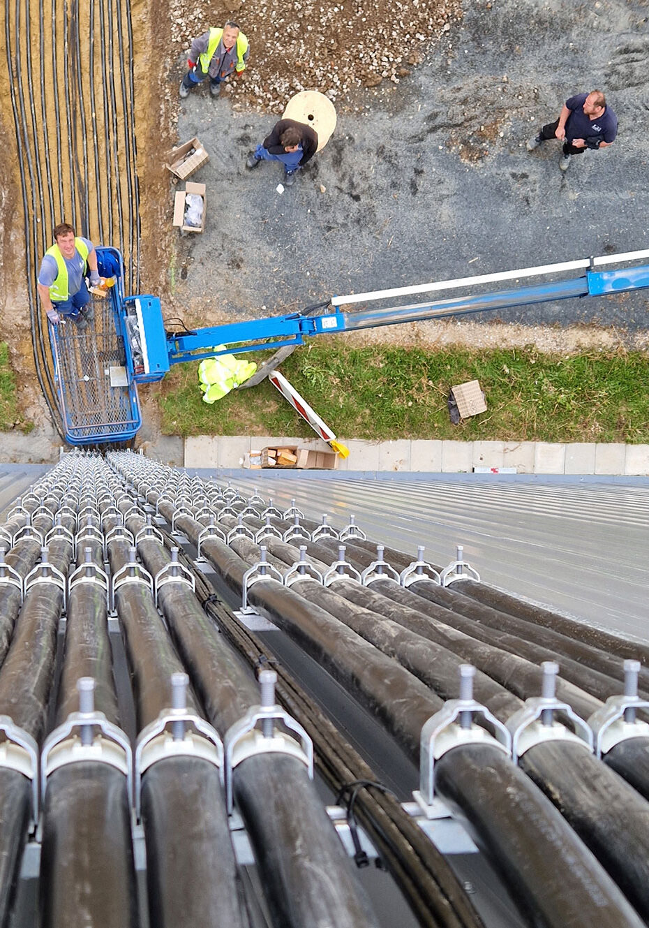 Blick von oben auf eine Baustelle mit Arbeitern, die an Kabeln und einer Hebebühne arbeiten, die entlang einer vertikalen Struktur verlegt sind.